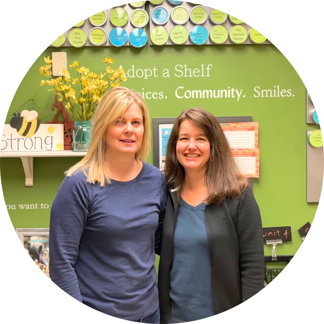 Two women smiling and standing in front of the green Adopt A Shelf wall in the FISH Marketplace.