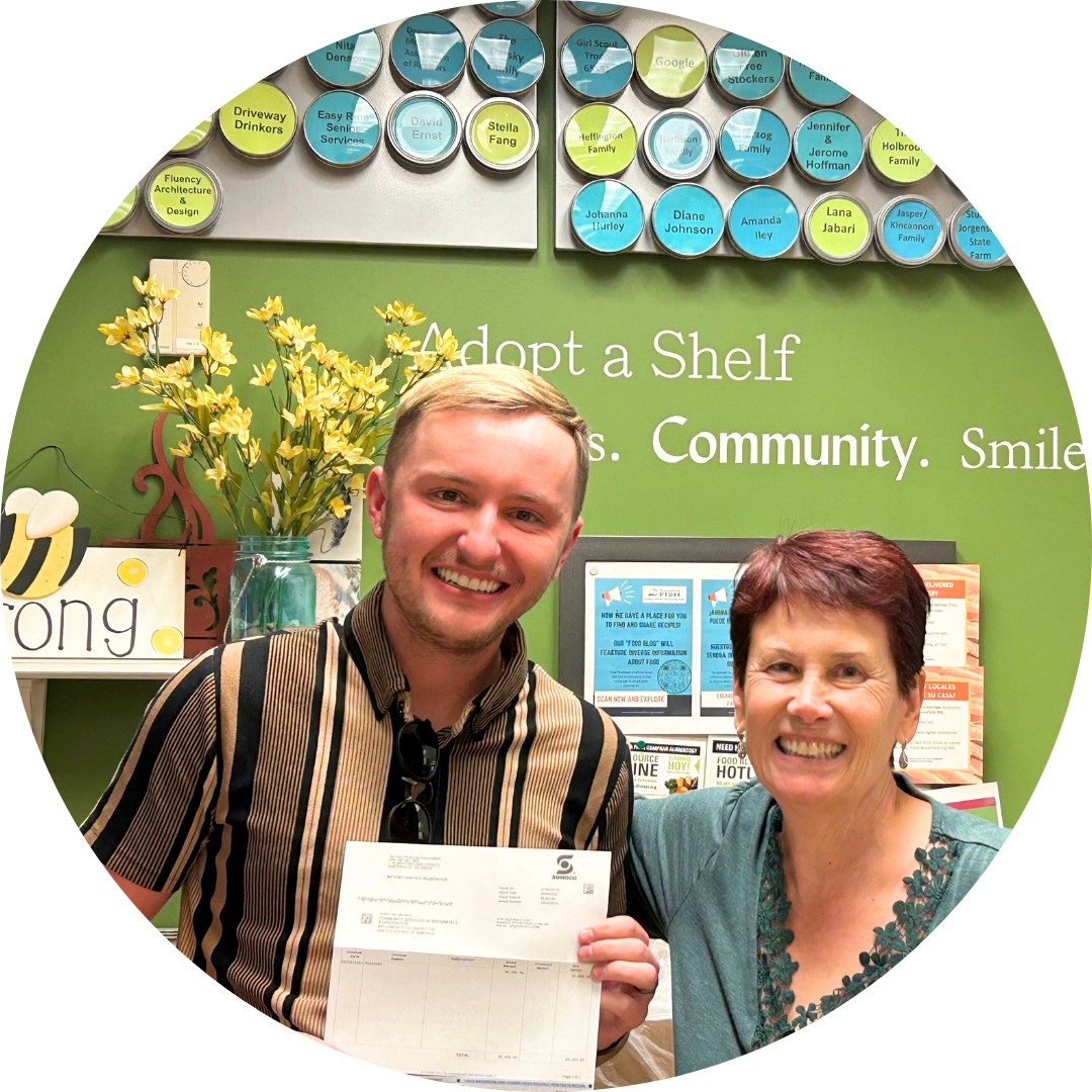 A man smiling while presenting a check donation to a FISH staff member in front of the green Adopt A Shelf wall in the FISH Marketplace.